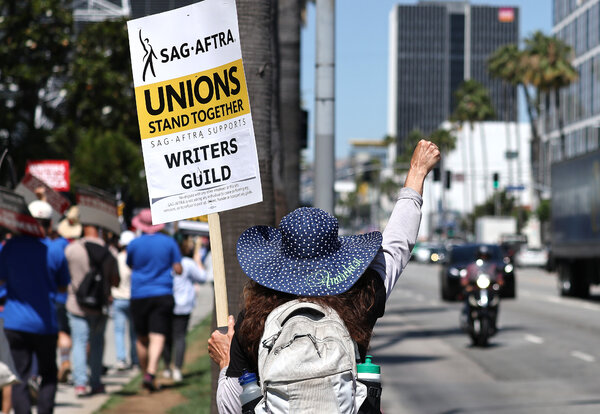 A street with protesters carrying signs. The closest one says “SAG-AFTRA Unions Stand  Together” and “SAG-AFTRA Supports Writers Guild.”